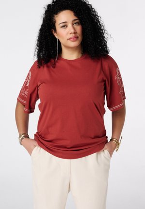Femme aux cheveux noirs bouclés porte un t-shirt rouge rouille avec une broderie blanche sur les manches et un pantalon crème, les mains dans les poches, fond neutre.