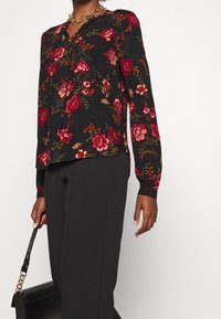 Blumenbluse aus schwarzem Stoff mit roten und grünen Mustern, langen Ärmeln und einem V-Ausschnitt. Kombiniert mit schwarzen Hosen und einer Handtasche.