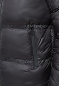 Giacca a vento nera con finitura lucida, cuciture orizzontali e tasca laterale con zip. Il materiale sembra morbido e isolato.