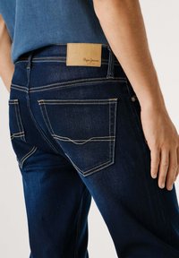 Tmavomodré denimové džínsy so rovnými nohavicami; obsahujú koženú nášivku s logom "Pepe Jeans" a svetlým prešívaním na okrajoch vreciek.