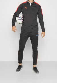 Schwarzer Sportanzug mit Reißverschlusskragen; rote Schulterpanels und blaues Logo auf der Hose. Schwarzer Fußball in der rechten Hand. Schwarze Schuhe.