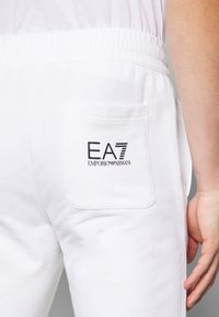 Bílé bavlněné joggingové kalhoty s zadní kapsou a černým logem EA7 Emporio Armani. Pas je elastický pro pohodlí.