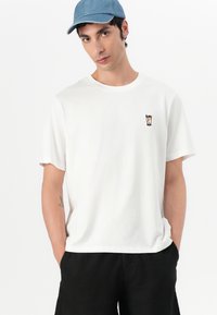 Hvit bomullst-skjorte med korte ermer, med et lite trykt grafisk motiv av en drink. Modellen har på seg en lyseblå caps og svarte shorts.