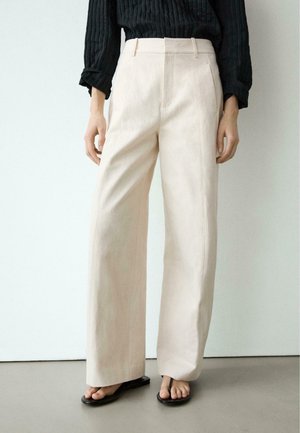 Donna che indossa pantaloni beige a vita alta e gamba larga abbinati a una maglia nera a costine a maniche lunghe e sandali neri infradito, in piedi contro un muro bianco.