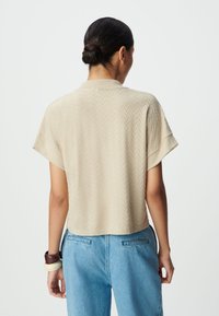 Krótki beżowy top z krótkim rękawem o teksturowanym wzorze w jodełkę, z płaskim kołnierzykiem i krótkim krojem, w zestawie z jasnoniebieskimi spodniami.