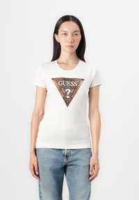 Hvid tætsiddende t-shirt med en trekantet brun grafik med teksten "GUESS?" og leopardmønstrede accenter, parret med blå denim jeans.