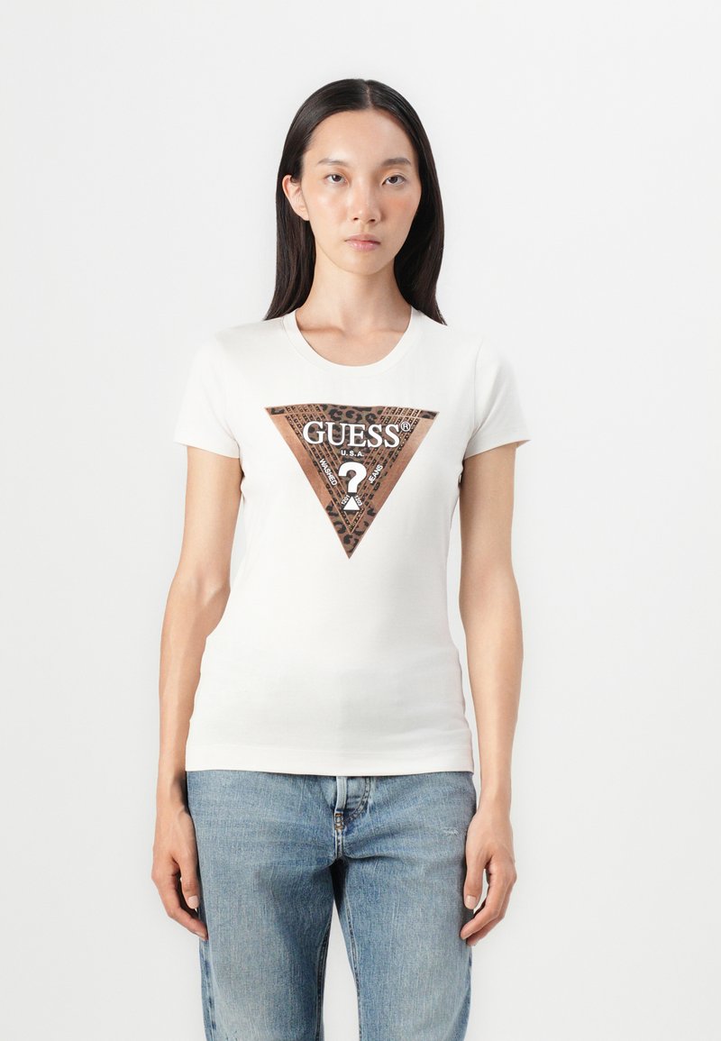 Hvid tætsiddende t-shirt med en trekantet brun grafik med teksten "GUESS?" og leopardmønstrede accenter, parret med blå denim jeans.