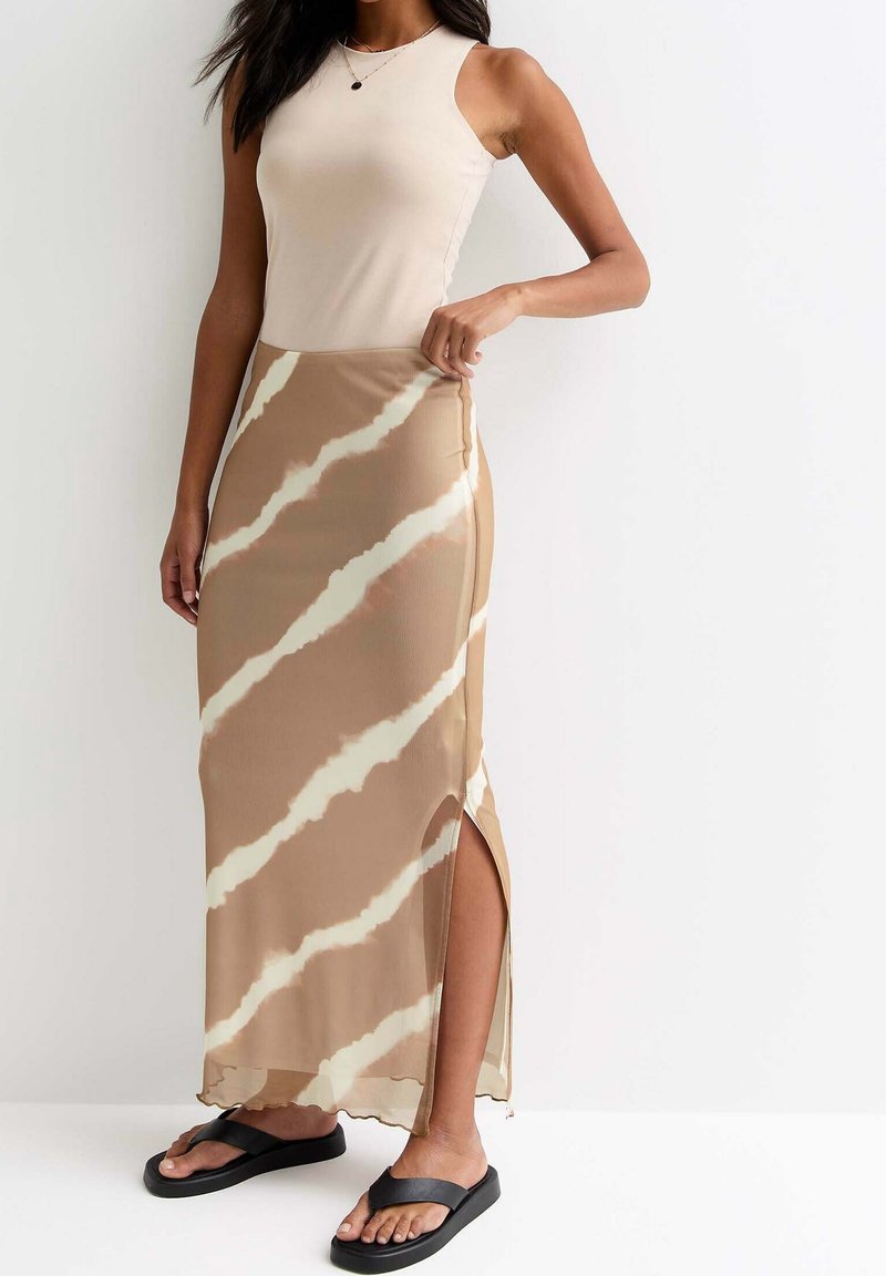 Femme portant un haut sans manches beige et une longue jupe beige et blanche à motifs avec une fente latérale et des sandales noires sur fond blanc.