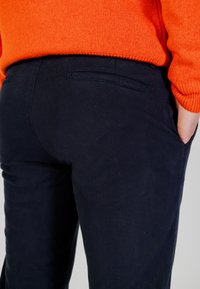 Pantaloni blu navy con tessuto strutturato, caratterizzati da due tasche sul retro e un design aderente, abbinati a un maglione a maglia arancione brillante.