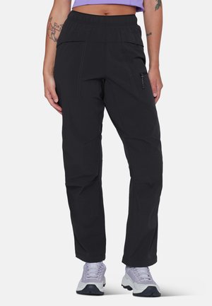 Donna che indossa pantaloni neri a gamba dritta con tasca laterale con zip e vita elasticizzata, abbinati a sneakers bianche e top viola corto.