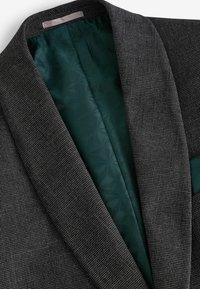 Blazer negro texturizado con un patrón de espiga, que presenta un interior de satén verde oscuro y un diseño de solapa elegante y estructurada.