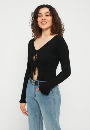 Γυναίκα με σγουρά μαλλιά που φοράει μαύρη κοντή μπλούζα με μακριά μανίκια και ριμπ ύφανση δεμένη στο μπροστινό μέρος, μαζί με ανοιχτόχρωμο τζιν ψηλόμεσο παντελόνι με μαύρη ζώνη.