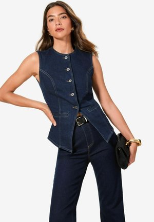 Dunkelblaue Denim-Weste ohne Ärmel mit fünf vorderen Knöpfen, schlanker Schnitt, seitlichen Taschen und Kontrastnähten. Getragen mit passenden Hosen und einer Clutch.