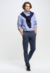 Camisa de botones de manga larga en azul claro; suéter azul marino drapeado sobre los hombros; pantalones ajustados oscuros en azul marino; zapatos negros; fondo neutro.