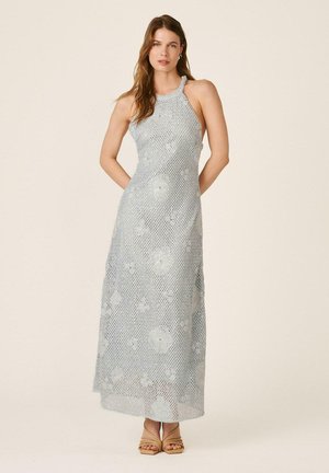 Mujer de pie vestida con un vestido largo sin mangas de ganchillo plateado grisáceo con detalles florales y sandalias de tacón beige.