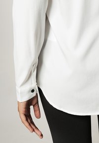 Blouse blanche à texture lisse, ourlet arrondi et poignets boutonnés. Le poignet est orné d'un bouton noir. Pantalon noir partiellement visible.