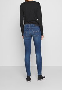 Jeans de mezclilla azul ajustados con bolsillos traseros que presentan un patrón de costura en forma de diamante, combinados con una camiseta negra de manga larga y zapatillas negras.