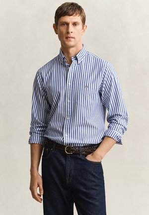 Mann trägt ein langärmliges, blau-weiß vertikal gestreiftes Hemd, das in eine dunkle Jeans mit braunem geflochtenem Gürtel gesteckt ist, steht mit einer Hand in der Tasche.