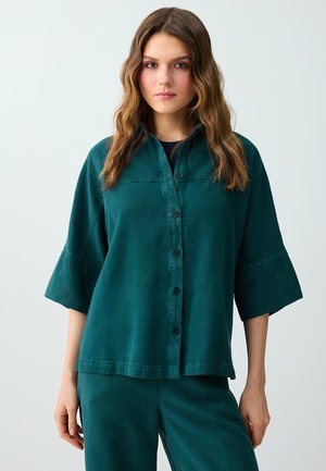 Femme aux longs cheveux ondulés porte une chemise turquoise boutonnée avec de larges manches et un pantalon assorti, debout devant un fond clair uni.
