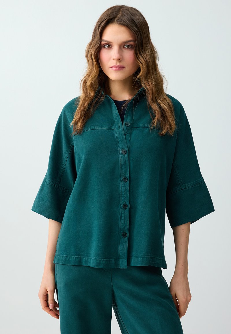 Femme aux longs cheveux ondulés porte une chemise turquoise boutonnée avec de larges manches et un pantalon assorti, debout devant un fond clair uni.
