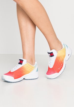 Persona con zapatillas blancas con lados en degradado de naranja a amarillo y acentos azules, de pie sobre un piso gris claro.