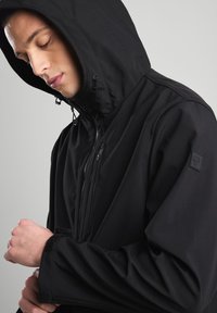 Black hooded jacket with a high collar, zippered front, and adjustable drawstrings. Soft texture, sleek design, featuring a logo patch on the sleeve.