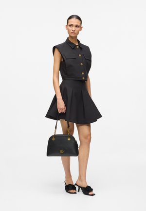 Woman wearing sleeveless black buttoned top and flared skirt, holding black handbag with gold chains, wearing black heeled sandals.