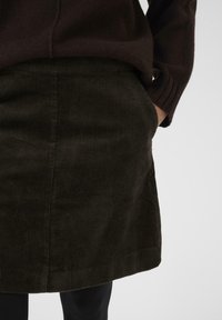 Une personne portant une jupe en velours côtelé marron foncé avec une main dans la poche, assortie à un pull à manches longues marron foncé, le tout sur un fond uni.