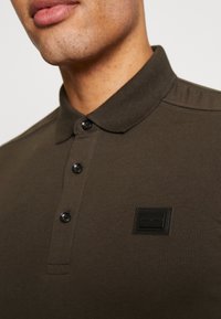Tmavě zelená polo košile s límečkem, čtyřmi černými knoflíky a obdélníkovou černou nášivkou na levé části hrudi. Měkká textura, štíhlý střih.