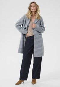 Donna che indossa un cappotto in tessuto a scacchi blu e beige, un top beige, pantaloni larghi scuri e stivali con tacco marroni, in piedi davanti a uno sfondo semplice.