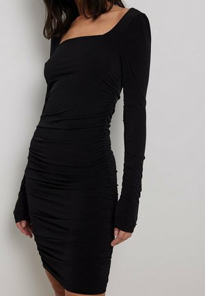 Kobieta w dopasowanej, czarnej sukience z marszczeniami, z długimi rękawami i kwadratowym dekoltem, stojąca na tle jasnego, jednolitego tła.