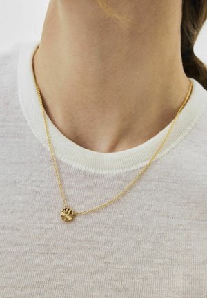 Artimojo asmens kaklo sritis, dėvinti ploną aukso grandinėlės vėrinį su maža plaktos aukso pakabuku ant balto tekstūruoto mezgimo viršutinio drabužio.