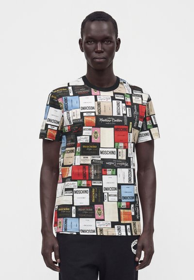 Άνδρας που φοράει ένα πολύχρωμο T-shirt Moschino με διάφορα σχέδια ετικετών, στέκεται μπροστά σε απλό φωτεινό φόντο.
