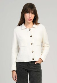 Femme portant un cardigan blanc à boutons avec un col et un jean noir, se tenant devant un fond gris clair uni.