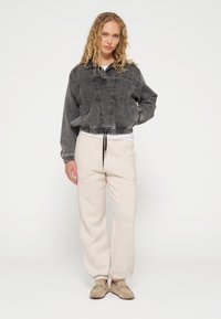 Giacca in denim nera con tasche frontali, orlo con cordino e maniche lunghe, abbinata a pantaloni della tuta beige chiaro e scarpe slip-on beige.