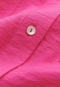 Tissu rose vif avec un gros plan d'un bouton blanc à deux trous cousu sur une couture. Le tissu présente un tissage texturé subtil.