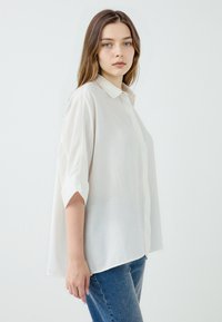 White, loose-fitting shirt with a collared neckline and rolled sleeves, made of lightweight fabric. Paired with blue high-waisted jeans.