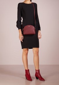 Schwarzes Kleid mit Puffärmeln, kombiniert mit einer burgunderfarbigen Leder-Crossbody-Tasche und roten Knöchelstiefeln mit spitzem Zehen und schrägem Blockabsatz.