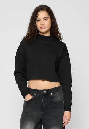 Sudadera negra recortada con cuello alto, ajuste holgado y mangas largas; combinada con vaqueros negros de talle alto con textura desgastada.