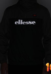 Černý lehký větrný plášť s kapucí, s reflexním logem "ellesse" přes hruď a elastickými manžetami na rukávech pro lepší přiléhavost.