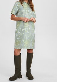 Kragskjortklänning med kort ärm i ljusblått med ett grönt blommönster; kombinerad med höga, mörkgröna gummistövlar.