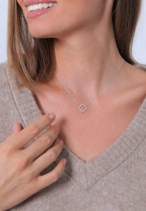 Silver necklace with a floral-shaped pendant featuring small clear stones, worn on a v-neck sweater, with a hand gently touching it.