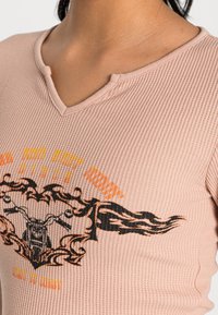 Svetle růžový žebrovaný top s výstřihem do V, který zdobí grafický potisk motocyklu s oranžovými a černými plamennými akcenty.