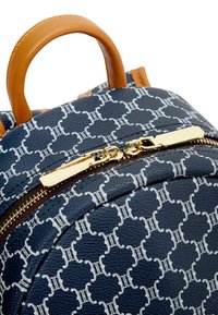 Marinblå ryggsäck med slät yta, guldfärgad dragkedjehårdvara och handtag i tan läder. Har ett upprepande vitt emblemdesign.