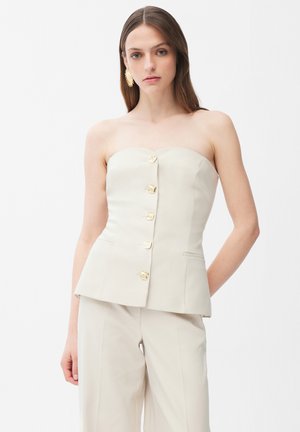 Kvinde iført en beige, stropfri top med fem gyldne knapper ned foran, kombineret med matchende beige bukser med vide ben.