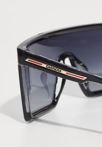 Occhiali da sole neri con montatura spessa, lenti grigie sfumate e branding accentato in oro. Presentano un design elegante con strisce rosse lungo la tempia.