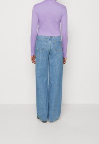 Osoba má na sobě světle modré džíny s vysokým pasem a širokými nohavicemi a na těle přiléhavý fialový top s dlouhými rukávy, stojí čelem k jednobarevné bílé zdi.