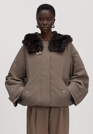 Femme portant une veste marron oversize avec un col en fausse fourrure noire et un pantalon marron ample assorti, debout devant un fond uni.