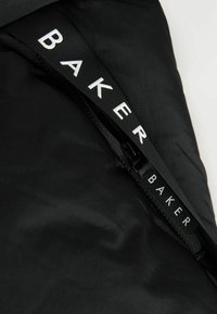 Zwarte stof met een gladde textuur heeft een rits en een riem met het label "BAKER" in vetgedrukte witte letters. Het ontwerp is minimalistisch en functioneel.