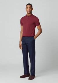 Bordeaux polo shirt met korte mouwen en een drieknops sluiting, gecombineerd met marineblauwe broek en bruine suède enkellaarzen. Effen achtergrond.